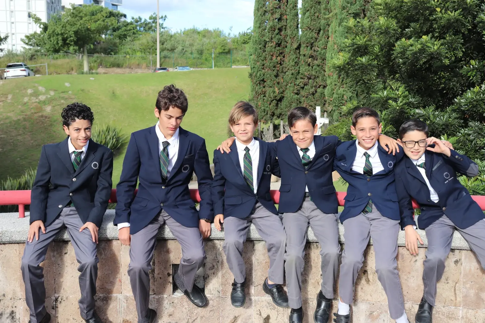 Seis estudiantes de uniforme escolar posan juntos en un banco de piedra en el campus, con áreas verdes al fondo