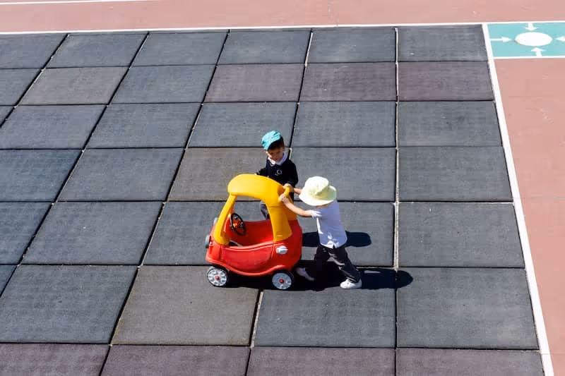 Dos niños jugando con un cochecito de juguete rojo y amarillo sobre baldosas de goma en una cancha.