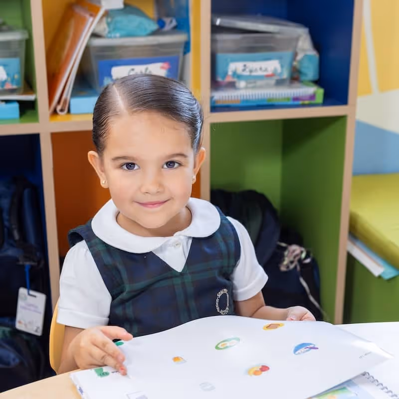 Niña con uniforme escolar sosteniendo papeles con dibujos de colores en un salón de clases con estantes al fondo.