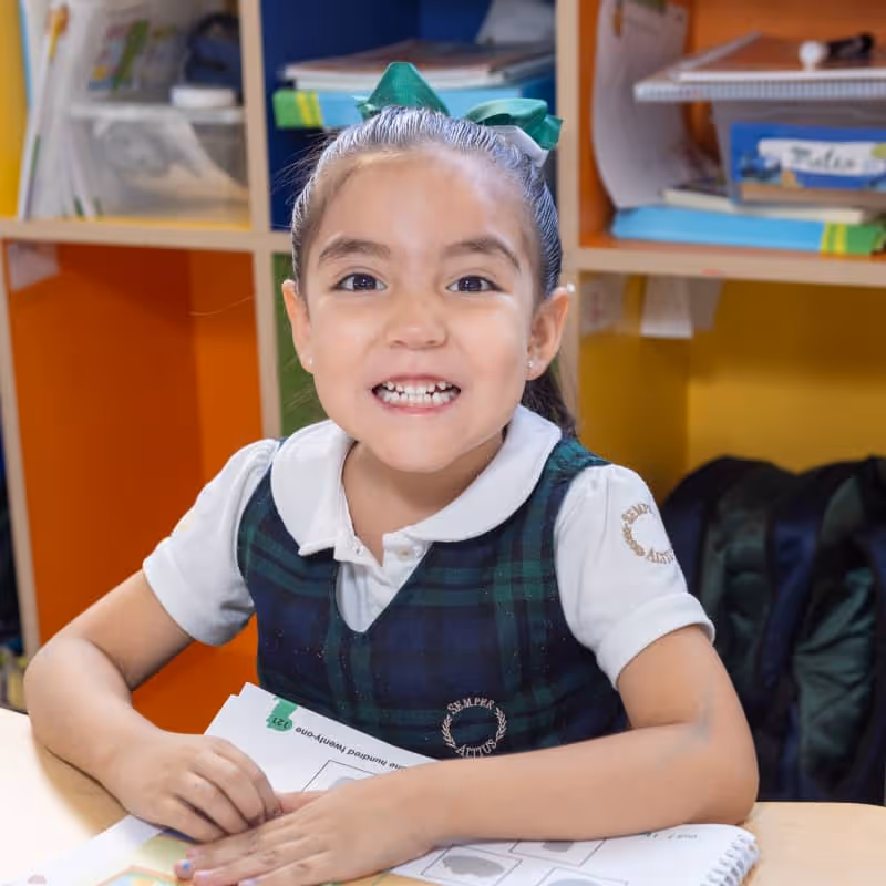 Niña sonriente con uniforme escolar sentado en un pupitre con libros y cuadernos en un salón de clases.