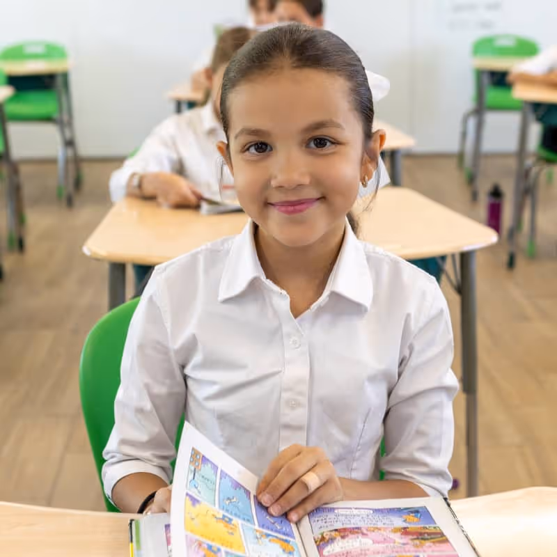 Niña sonriente en uniforme blanco sentada en un aula con libro abierto de historietas.