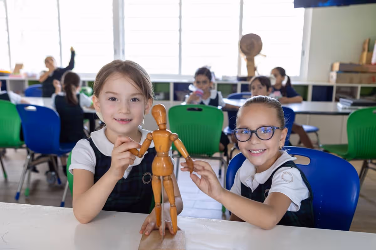 Dos niñas en uniforme escolar sonríen mientras sostienen una figura de madera articulada en un salón de clases con sillas verdes y azules.